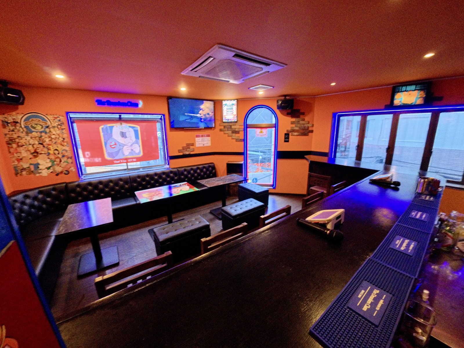 Interior of The Drunken Clam, a lively open-mic karaoke bar in Osaka near Dotonbori, with a long bar counter, lounge seating, colorful neon lighting, TVs, and karaoke microphones.